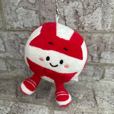 A red and white striped plush character keychain with a smiling face and small legs, hanging from a metal chain against a gray stone wall.
